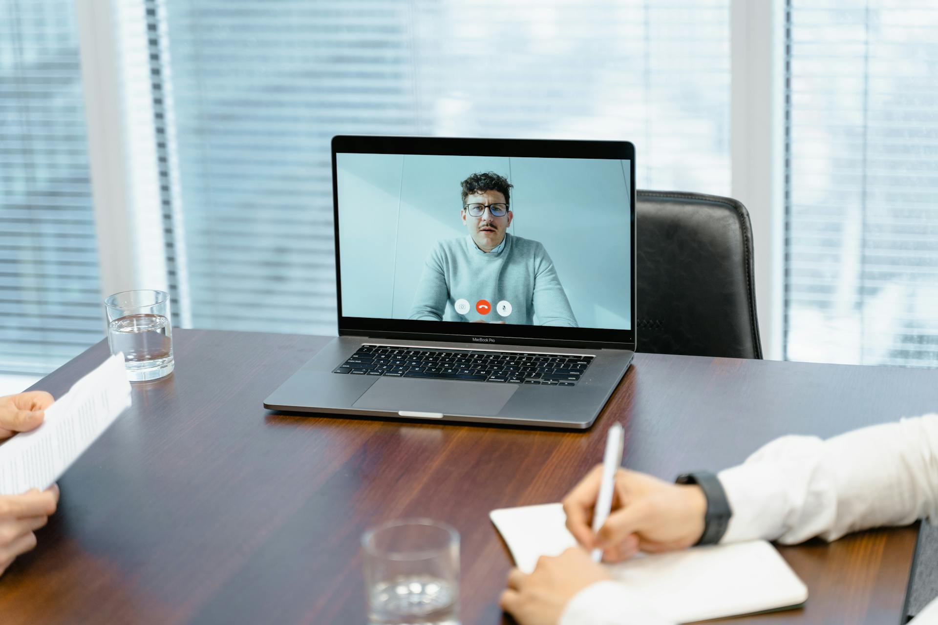 A laptop on a conference table shows a man participating in a video call during remote deposition, while another person in the foreground takes notes.