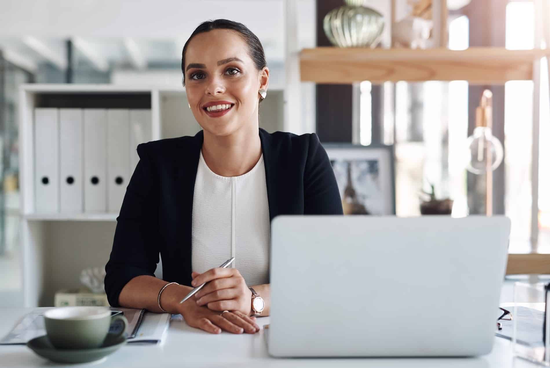 A smiling female professional sitting at a desk in a modern office with a laptop, pen, and coffee cup.